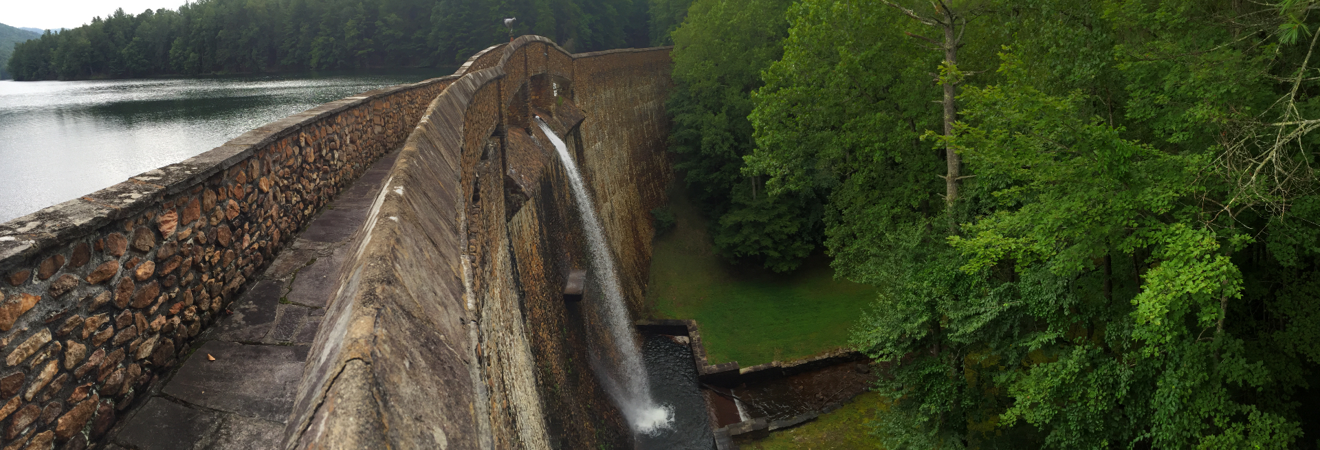Panorama along the stone crest walkway of the Devotion hydroelectric dam, the lake on the left and the spillway falling into the green gorge on the right